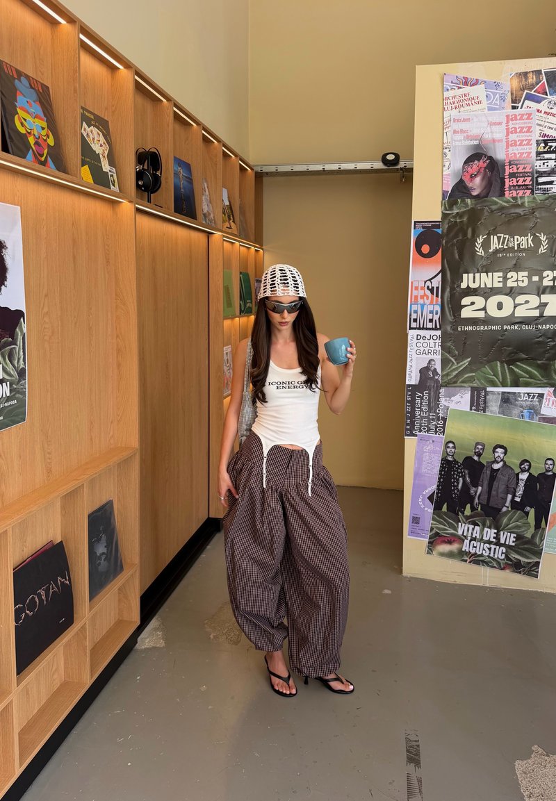 Femme portant des lunettes de soleil et un pantalon large à carreaux tient une tasse bleue dans une pièce avec des étagères en bois et des affiches de festival de jazz au mur.