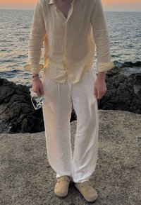 Ανοιχτό κίτρινο λινό πουκάμισο, λευκά λινά παντελόνια, μπεζ παντόφλες. Το άτομο κρατάει γυαλιά ηλίου, στέκεται σε βραχώδη επιφάνεια κοντά στη θάλασσα.