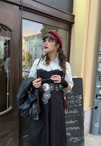 Čierne maxi šaty s čipkovým prekrytím, zdobené kovovými hrotmi a veľkou sponou na opasku. Biely dres pod nimi, kožená bunda a červený baret.