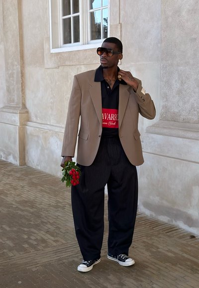 Hombre con blazer beige de gran tamaño, pantalones negros, gafas de sol, sosteniendo flores rojas, de pie en interiores junto a una pared con textura y una ventana.