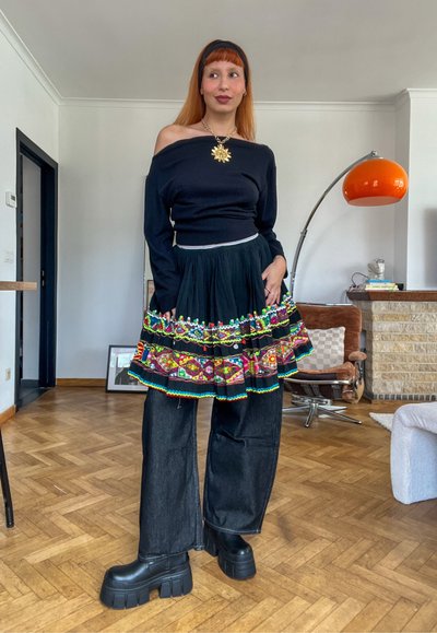 Mujer con cabello rojo que lleva una blusa negra sin hombros, falda bordada colorida sobre pantalones anchos negros y zapatos negros gruesos de plataforma en interiores.