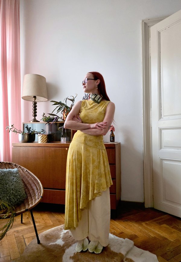 Γυναίκα με γυαλιά, σε κίτρινο φόρεμα και μπεζ παντελόνι, όρθια με σταυρωμένα χέρια σε ένα σαλόνι με ξύλινα έπιπλα και φυτά.