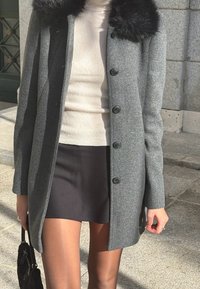 Nő szürke, gombos kabátot visel fekete szőrme gallérral, bézs színű bordázott pulóverrel, fekete szoknyával, átlátszó harisnyával, és fekete kézitáskát tart a kezében.