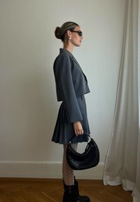 Blazer cinzento curto e saia plissada, combinados com uma mala trançada preta. Meias-calças transparentes com padrão e botas pretas de tornozelo completam o visual.