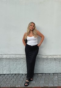 Svart midikjol med hvite prikkar, smal passform, høyt liv, kombinert med ein kvit singlet og svarte sandalar. Modellen står mot ei lys vegg.