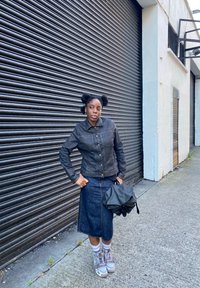 Giacca di jeans nera con bottoni dorati, gonna di jeans blu, sneakers grigio chiaro e viola, che tiene una borsa nera, in piedi contro un muro strutturato.