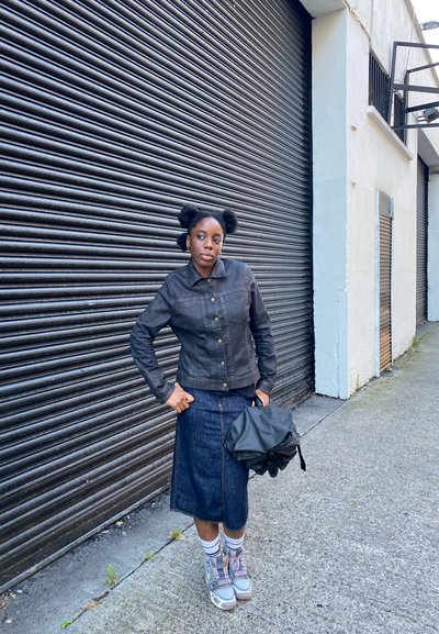 Chaqueta de denim negra con botones dorados, falda de denim azul, zapatillas de deporte en gris claro y púrpura, sosteniendo un bolso negro, de pie frente a una pared texturizada.