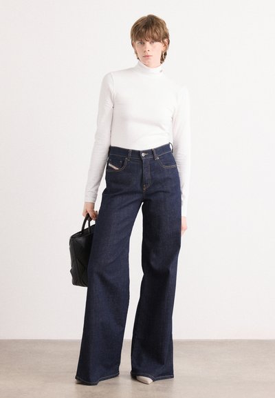 Pantalones vaqueros de pierna ancha azul oscuro, confeccionados en denim, con cintura alta, bolsillos delanteros y costuras doradas. Llevados con un cuello de tortuga blanco ajustado.