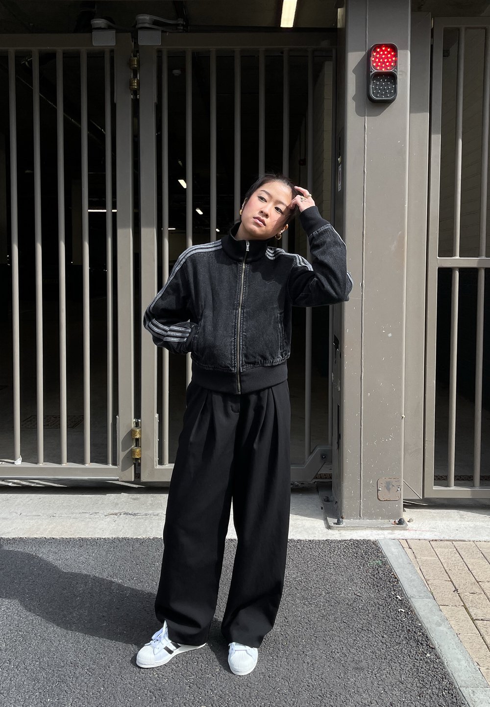 Casaco preto cropped com colarinho alto, combinado com calças largas pretas e sapatilhas brancas. O fundo apresenta uma área com portão e uma luz vermelha.