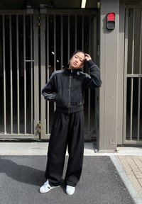 Casaco preto cropped com colarinho alto, combinado com calças largas pretas e sapatilhas brancas. O fundo apresenta uma área com portão e uma luz vermelha.