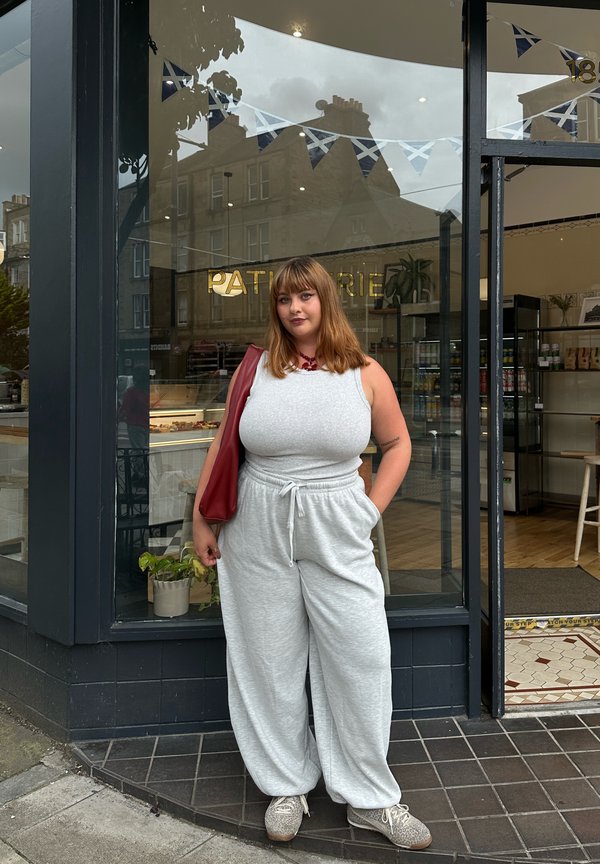 Jeune femme en haut gris sans manches et pantalon se tient devant une pâtisserie avec une guirlande de drapeaux écossais visible à l'intérieur.