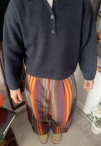 Black knitted sweater with three buttons, paired with pleated trousers featuring multicoloured horizontal stripes in red, green, and purple, completed with beige shoes.