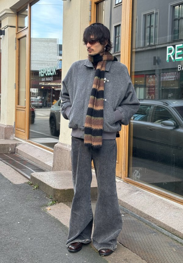 Gray oversized jacket with button accents, paired with wide gray pants. Black and brown striped scarf. Brown shoes. Urban outdoor setting.