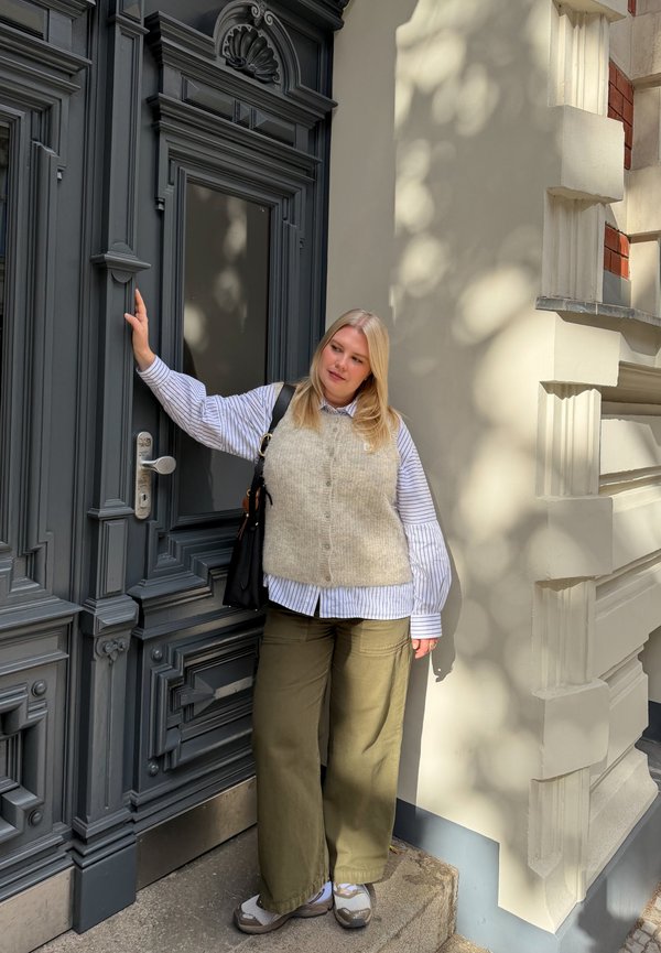 Jeune femme aux cheveux blonds portant un gilet gris, une chemise rayée et un pantalon vert se tient près d'une porte sombre ornée dans une rue ensoleillée.