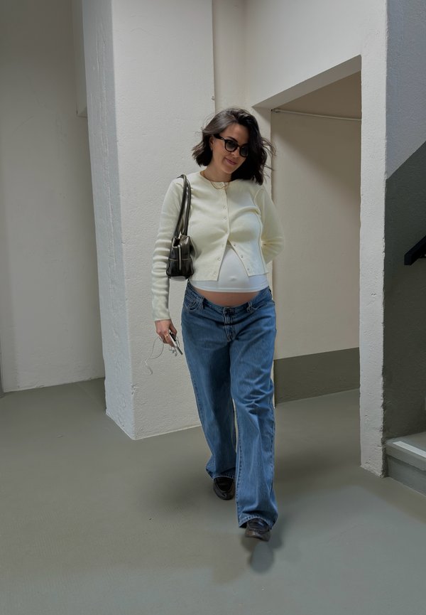 Gravid kvinne iført solbriller, hvit cardigan og blå jeans som går innendørs nær hvite vegger og trapper, holder en veske og nøkler.