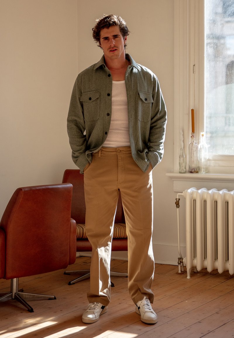 Pilkas marškiniai su dviem priekinėmis kišenėmis, balta palaidinė, smėlio spalvos kelnės ir baltos sportiniai bateliai, stovintys viduje, prieš minimalų foną.