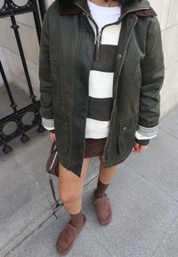 Olivgrüne gewachste Jacke mit einem Cordkragen, gestreiftem Pullover, brauem Minirock, braunen Socken und Wildlederschlappen. Auffällige Taschen Details.