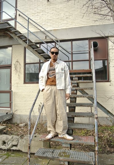 Chaqueta bomber blanca, camiseta gráfica marrón de Nike, pantalones beige oversize y zapatillas blancas. Escalera de metal y pared de ladrillo de fondo.