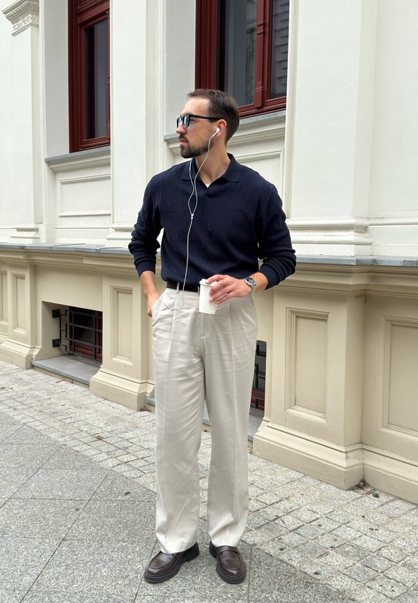Hombre con suéter oscuro, pantalones claros, gafas de sol y auriculares, de pie en la acera sosteniendo una taza de café blanca junto a un edificio beige.