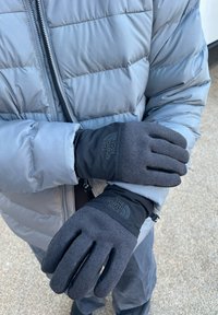 Luvas pretas feitas de um tecido suave e texturizado, com punho e destaque de logotipo. Usadas por uma pessoa com uma parka cinza.