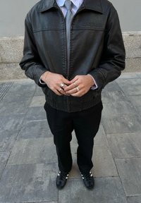 Casaco bomber de couro preto com bainha canelada, usado sobre uma camisa listrada clara e uma gravata cinza. Os acessórios incluem vários anéis e calças pretas.