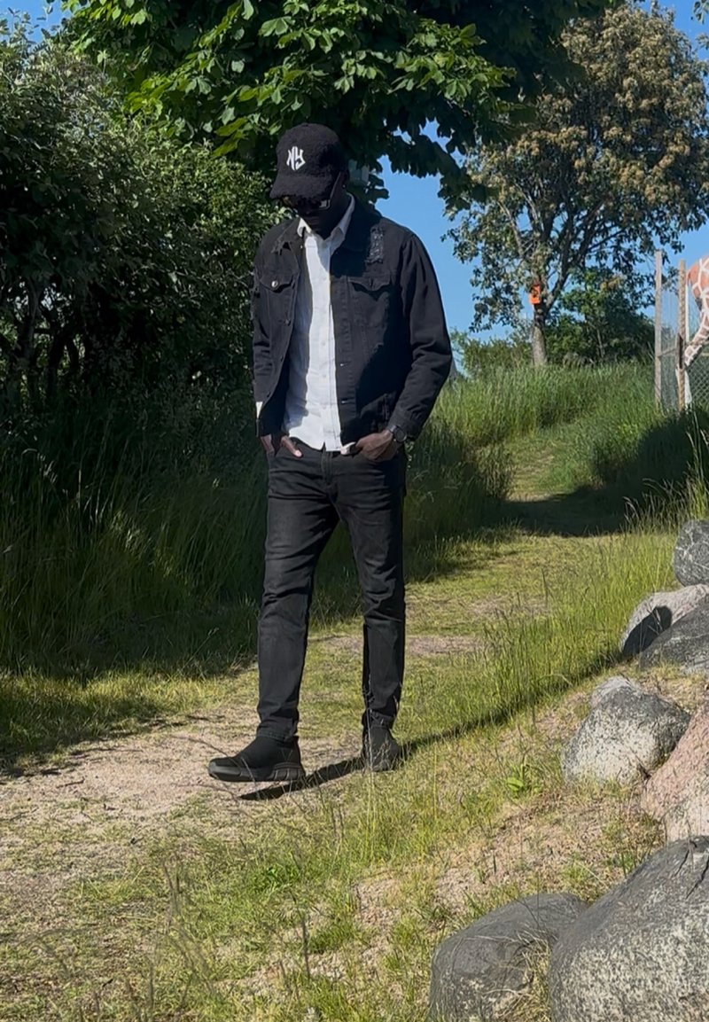 Homme portant une veste noire, une chemise blanche et une casquette noire marchant sur un sentier herbeux baigné de soleil entouré d'arbres et de rochers.