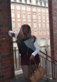 Dzianinowy sweter z geometrycznym wzorem w odcieniach szarości, czerni i bieli; bordowa spódnica; brązowa torebka; w tle ceglana i szklana architektura.