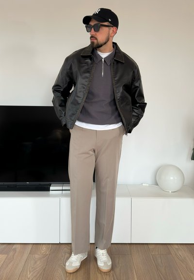 Chaqueta de cuero negra, suéter gris con cuello y cremallera, pantalones beiges y zapatillas blancas con detalles grises. Gafas de sol oscuras y gorra.