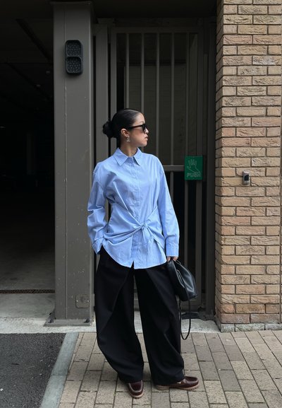 Mujer con gafas de sol que lleva una camisa azul abotonada anudada a la cintura, pantalones negros anchos y sostiene un bolso negro cerca de una pared de ladrillos y una puerta.