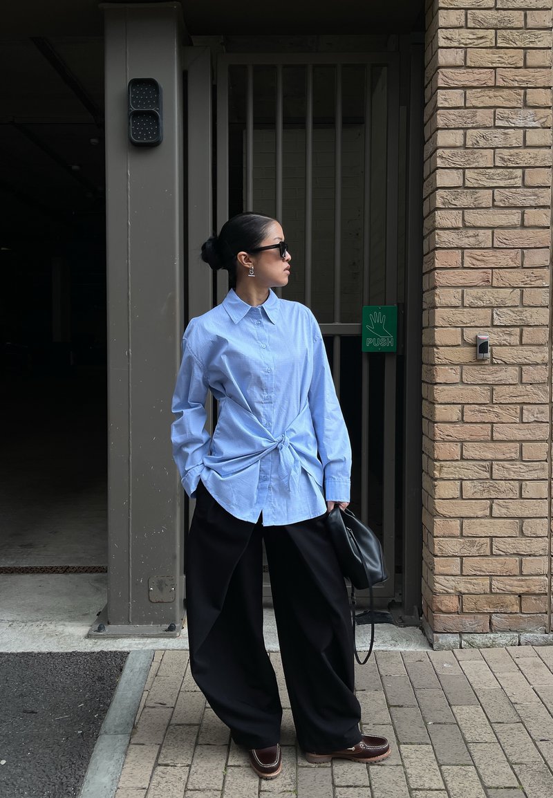 Frau mit Sonnenbrille, die ein blaues Hemd mit Knöpfen trägt, das in der Taille gebunden ist, weite schwarze Hose und eine schwarze Tasche hält, neben einer Ziegelmauer und einem Tor.