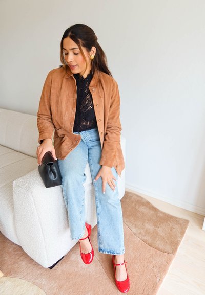 Mujer sentada en un banco blanco texturizado con chaqueta marrón, top negro de encaje, vaqueros azul claro, tacones rojos de charol, sosteniendo un bolso de mano negro.