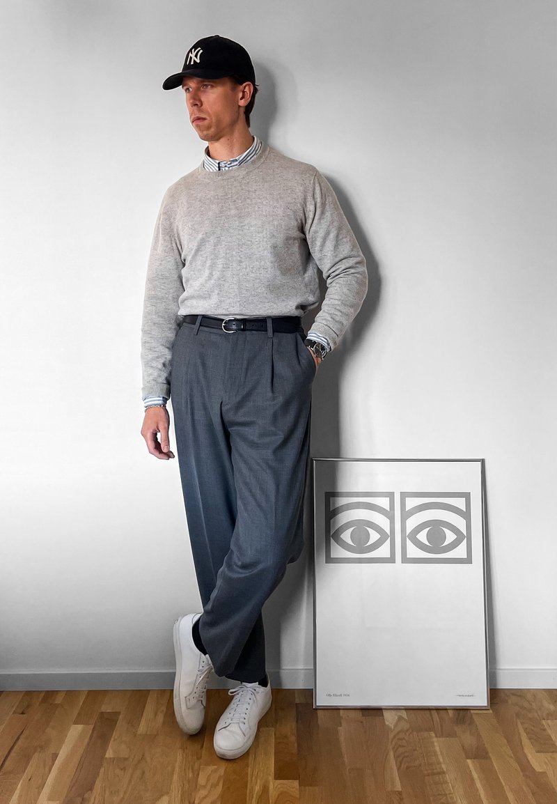 A grey jumper over a blue-and-white striped shirt, grey pleated trousers, white sneakers, and a black cap; an art print featuring eyes in the background.