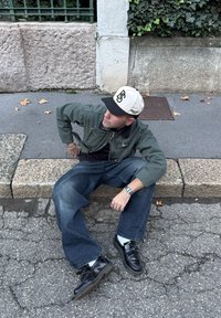 Um homem senta-se num meio-fio de pedra, vestindo uma jaqueta bomber verde, jeans azuis, sapatos pretos e um boné bege com letras pretas.