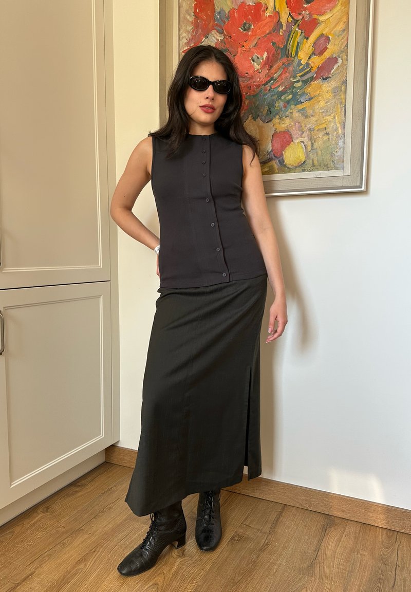 Femme portant un haut noir sans manches, une longue jupe noire, des bottes noires et des lunettes de soleil foncées, debout à l'intérieur près d'une peinture florale colorée.