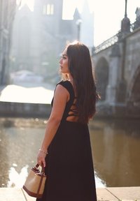 Sort ærmeløs kjole med en rund udskæring og udskåret rygdesign. Modellen holder en vævet skuldertaske med et læderhåndtag. Baggrunden har en flod og en bro.