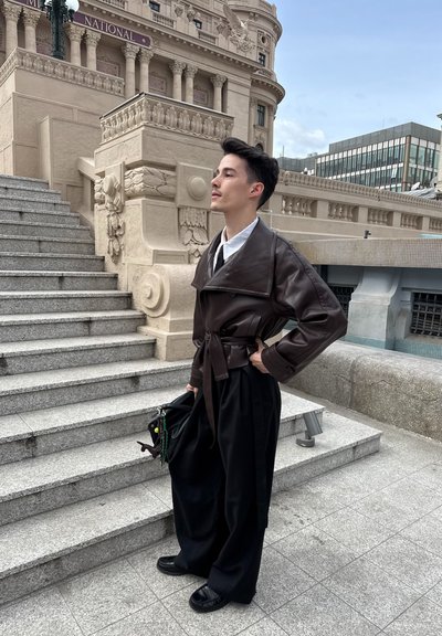 Persona joven con chaqueta de cuero oscura y pantalones anchos negros está cerca de unos escalones de piedra y una fachada de edificio ornamentada, sosteniendo una bolsa negra.