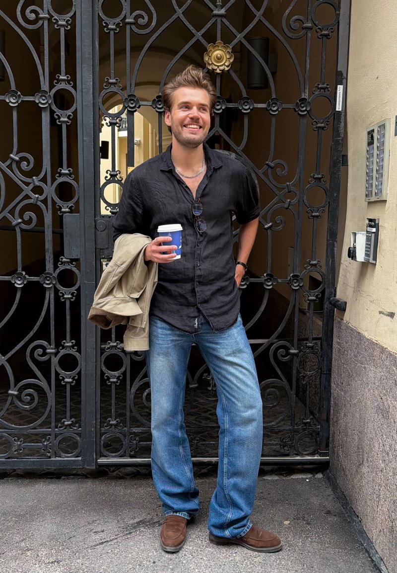 Ung mann smiler, holder en kaffekopp og en beige jakke, står foran dekorative svarte smijernsporter i en bygate.