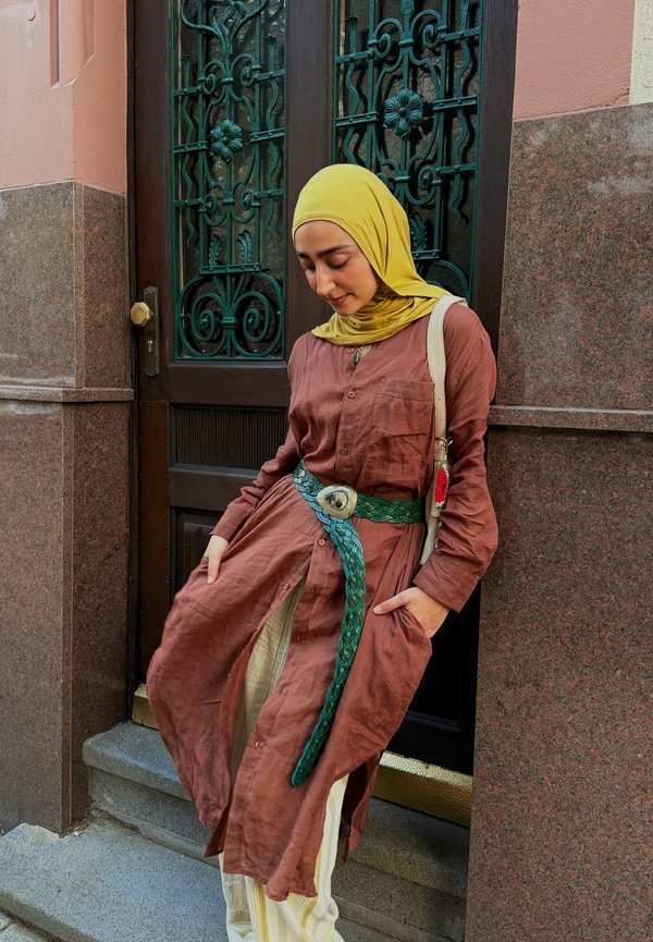 Woman wearing a yellow headscarf and brown dress with green belt, standing on steps in front of a decorative dark wooden door.