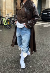 Personne portant un manteau en cuir marron avec col en fourrure, chemise à carreaux marron raccourcie, jean ample bleu clair et baskets blanches montantes, marchant sur un trottoir mouillé.