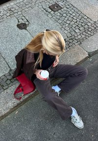 Kvinna med blont hår och brun jacka sitter på en kantsten och håller i en vit kopp, röd handväska bredvid sig, klädd i mörka byxor och vita sneakers.