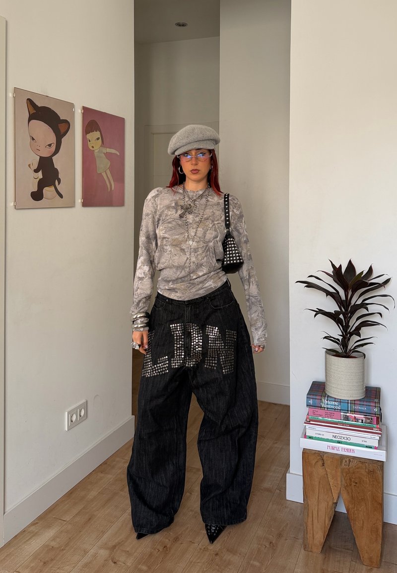 Person wearing gray patterned top, gray beret, oversized black studded pants with "LJDN," standing in room with two wall art pieces and potted plant.