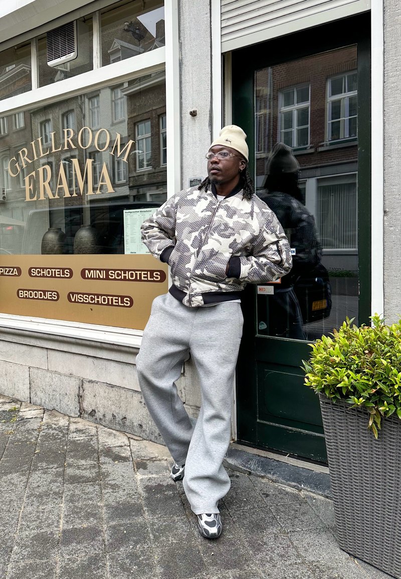 Kamouflage-mönstrad bomberjacka, grå joggingbyxor, grå sneakers. Svart bågar glasögon, krämfärgad mössa, står mot en butiksvittrappa.