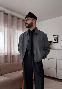 Homme avec barbe et lunettes porte un bonnet noir, une veste grise, une chemise noire et une cravate, debout dans un salon avec des rideaux, un canapé et un meuble de rangement.