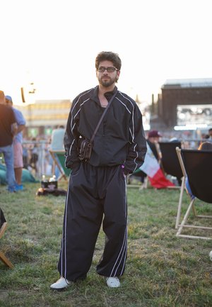 Schwarzer Jogginganzug mit weißen Streifen, übergroßer Schnitt, getragen mit einer kleinen braunen Umhängetasche und weißen Schuhen, stehend auf Gras bei einer Freiluftveranstaltung.