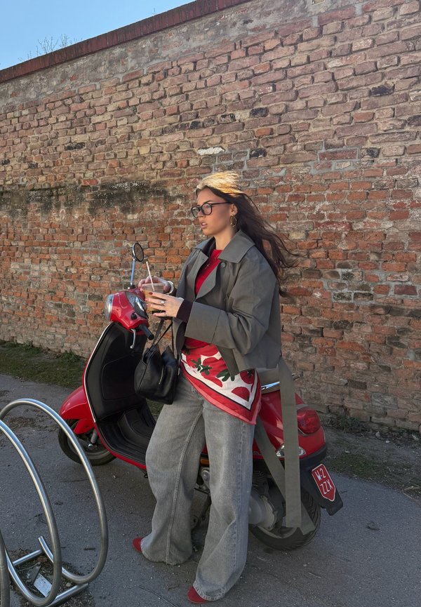 Femeie cu ochelari, purtând o geacă gri și un top floral, stă lângă un scuter roșu, ținând o băutură lângă un zid de cărămidă, sub un cer senin.