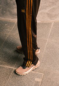 Brown athletic trousers with orange stripes, teamed with beige trainers that have a textured upper and black accents, positioned on a grey floor.