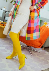 Personne portant un manteau à carreaux coloré sur une robe blanche avec des collants jaune vif et des chaussures à talons assorties, tenant un sac à main beige.