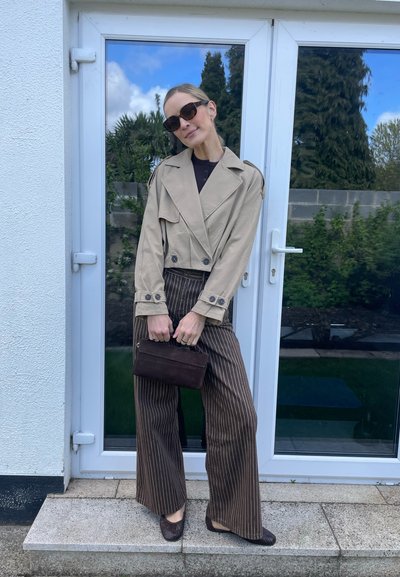 Mujer con chaqueta corta beige, pantalones anchos de raya marrón, gafas de sol oscuras, sujetando un bolso pequeño oscuro, de pie junto a una puerta de cristal al aire libre.
