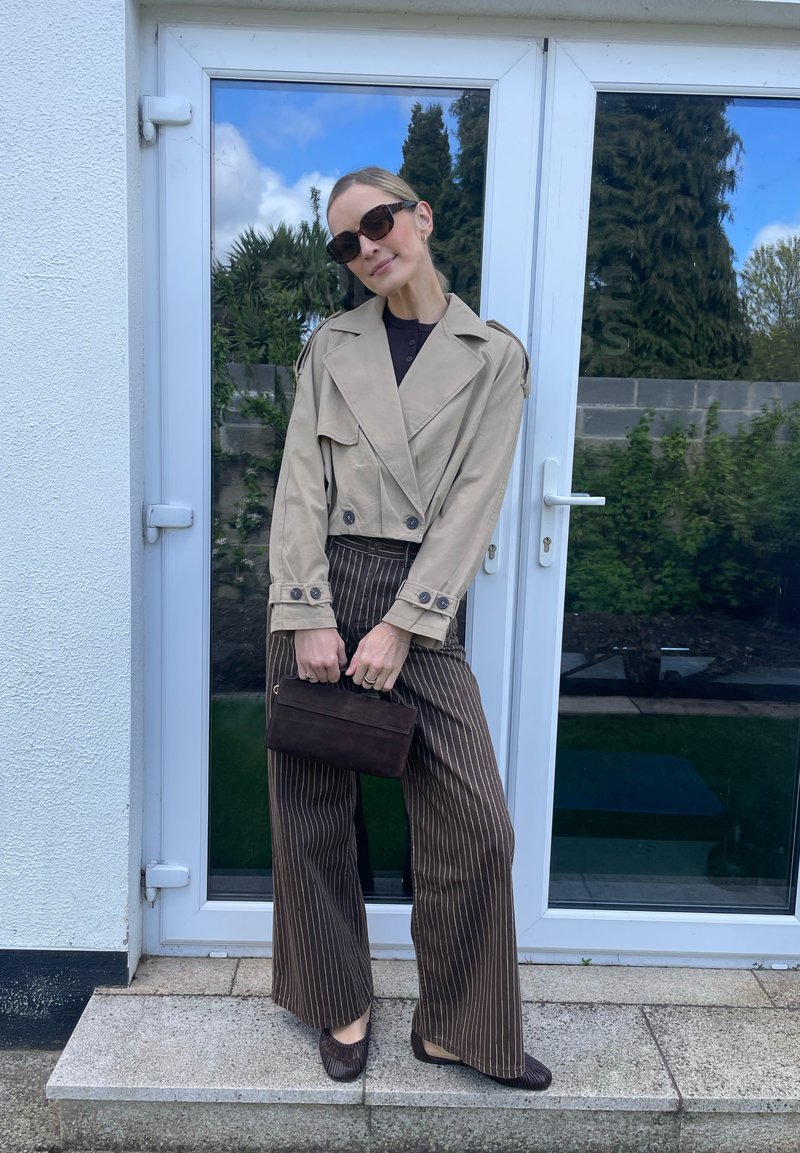Femme portant une veste courte beige, pantalon large marron à fines rayures, lunettes de soleil foncées, tenant un petit sac à main sombre, debout près d'une porte en verre en extérieur.