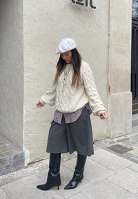 Pullover in crema a trecce, camicia a quadri sotto, culotte grigie e stivali neri al ginocchio; indossa un berretto bianco. Sfondo di marciapiede.